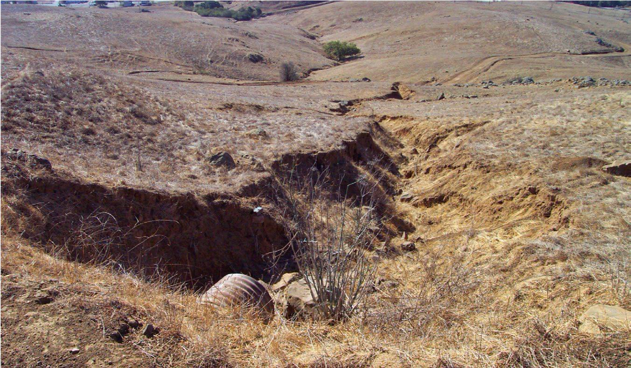 On a brown, grassy landscape, a long, jagged-edged gully is shown at the outlet of a culvert.