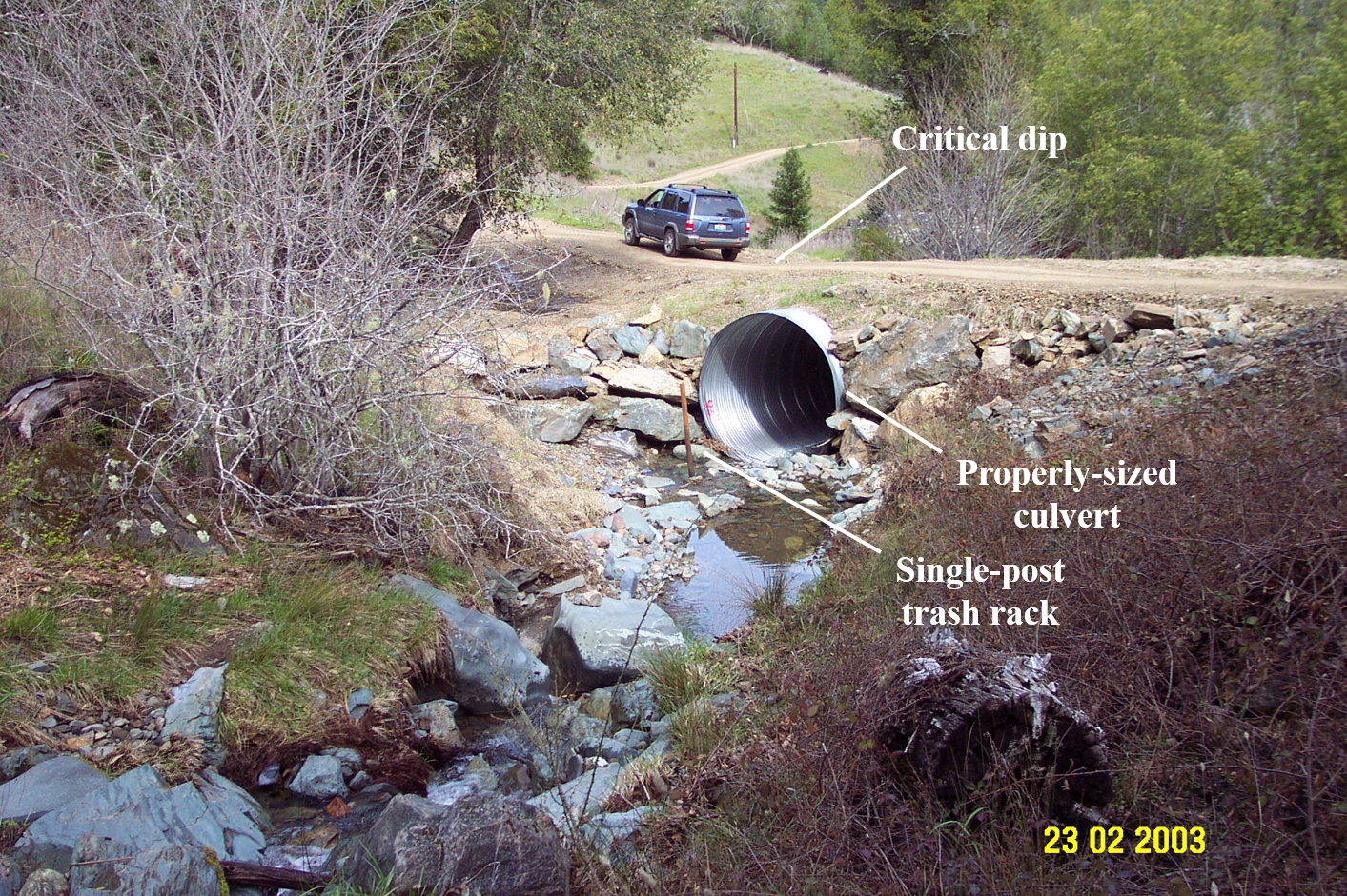 A larger view of a dirt road shows a culvert (with an associated trash rack). About 5-10 feet up from the culvert, a critical dip (a literal dip in the road) has been installed to ensure the stream is not diverted in a storm