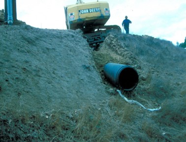 A backhoe and a person are shown with a half-buried culvert, demonstrating the angle of install.  