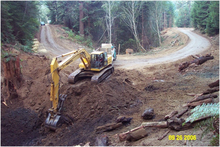  In this image, the same site has been cleared of vegetation. The road surface looks smooth, and a back-hoe is building up the bank on the side of the road.  