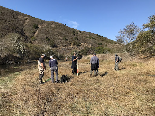 In the same location as the first image, 6 people are gathered, seeming to discuss the landscape. One is dressed in waders and another has a measuring device.